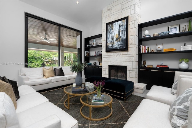 living room with a stone fireplace, built in features, and ceiling fan