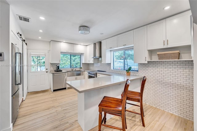 kitchen with a kitchen bar, appliances with stainless steel finishes, wall chimney range hood, a barn door, and white cabinetry