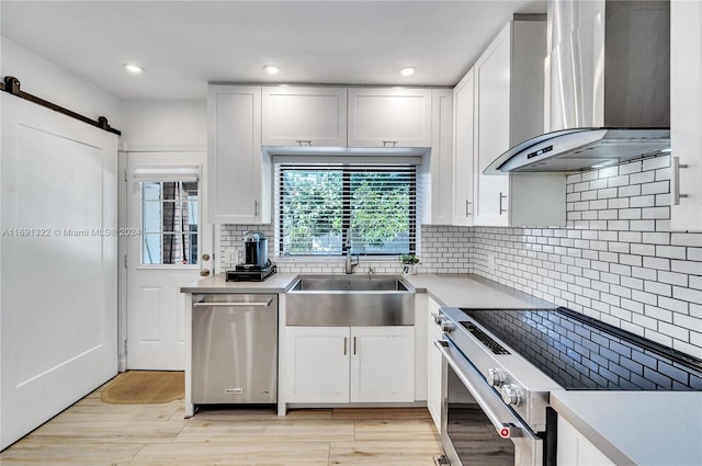 kitchen featuring appliances with stainless steel finishes, sink, wall chimney range hood, a barn door, and white cabinets