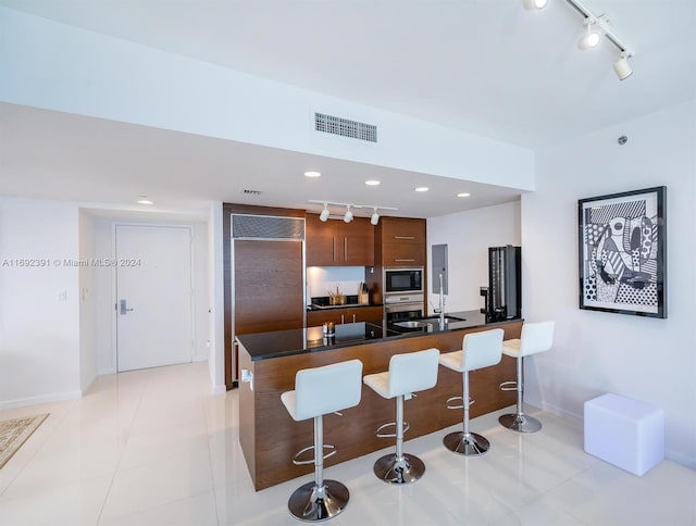 kitchen with light tile patterned floors, built in appliances, sink, rail lighting, and a breakfast bar area