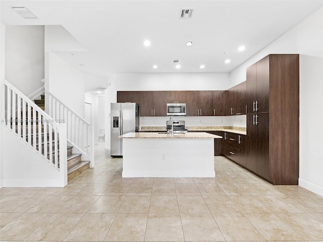 kitchen featuring sink, a kitchen island with sink, dark brown cabinets, stainless steel appliances, and light stone countertops