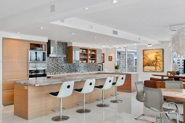 kitchen featuring a spacious island, decorative backsplash, wall chimney range hood, oven, and a kitchen bar