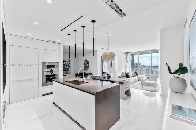 kitchen featuring white cabinets, sink, floor to ceiling windows, a kitchen island with sink, and pendant lighting