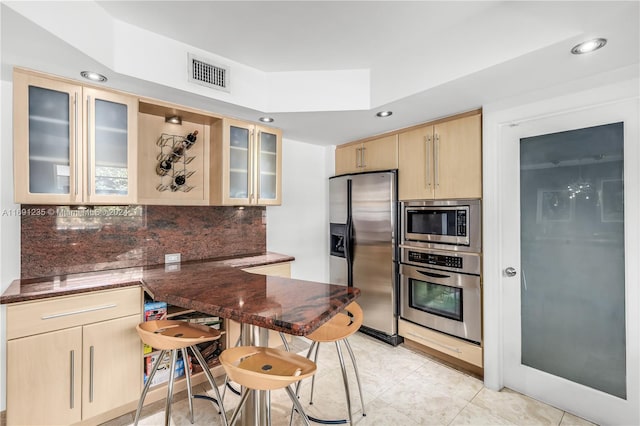 kitchen featuring dark stone countertops, light tile patterned floors, light brown cabinetry, appliances with stainless steel finishes, and kitchen peninsula