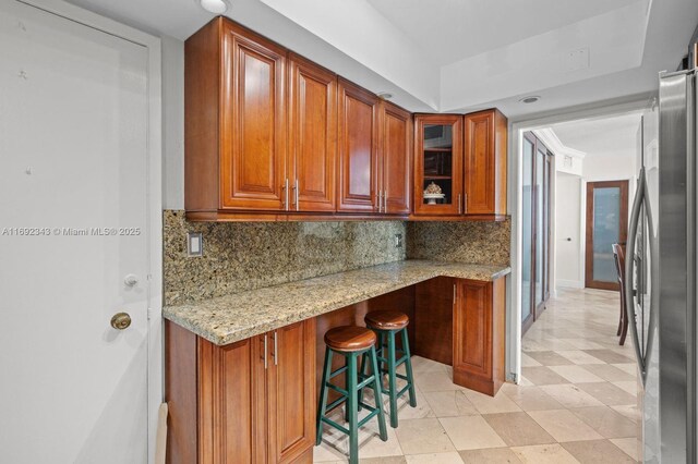 kitchen with light stone counters, a kitchen bar, backsplash, and stainless steel refrigerator