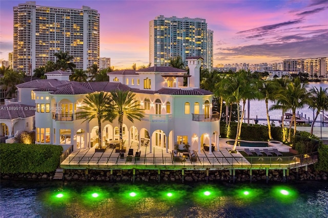 back house at dusk with a patio area, a balcony, a water view, and a pool