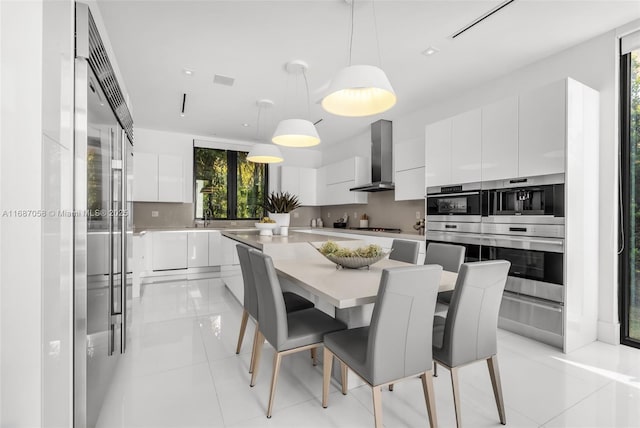 interior space featuring wall chimney range hood, white cabinetry, hanging light fixtures, a center island, and tasteful backsplash