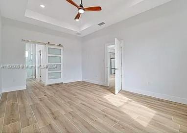 unfurnished room with ceiling fan, a barn door, a tray ceiling, and light hardwood / wood-style flooring