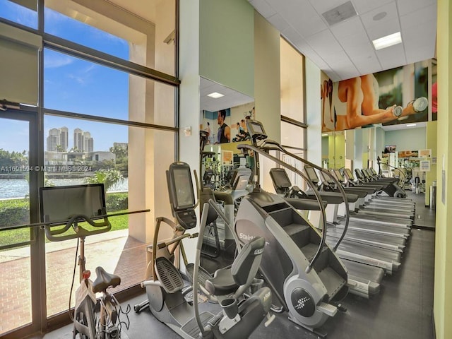 exercise room featuring a paneled ceiling, a water view, a wall of windows, and a towering ceiling