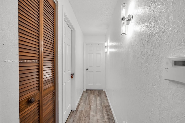 corridor featuring baseboards, a textured wall, light wood-type flooring, and a textured ceiling