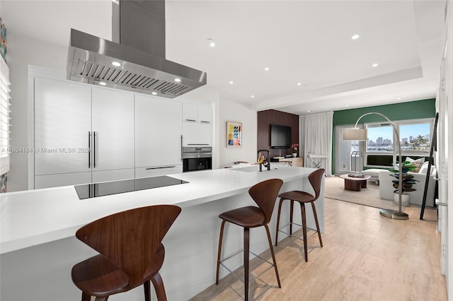 kitchen featuring black electric stovetop, stainless steel oven, a breakfast bar, wall chimney range hood, and white cabinetry