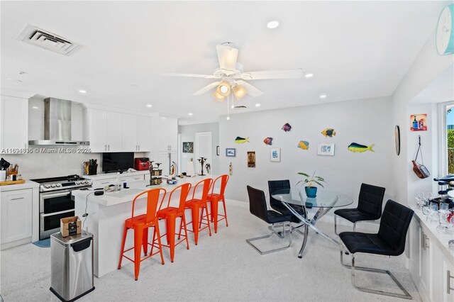 kitchen with a kitchen breakfast bar, wall chimney exhaust hood, ceiling fan, double oven range, and white cabinetry