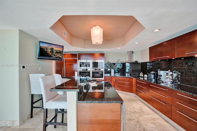 kitchen featuring decorative backsplash, a tray ceiling, a kitchen island with sink, wall chimney range hood, and a breakfast bar area