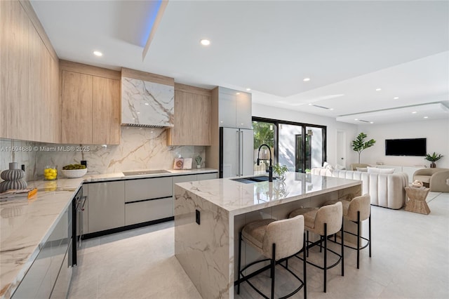 kitchen with sink, a breakfast bar area, a kitchen island with sink, light stone countertops, and black electric cooktop