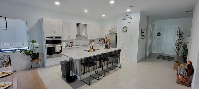 kitchen featuring a breakfast bar, white cabinets, wall chimney exhaust hood, an island with sink, and stainless steel appliances