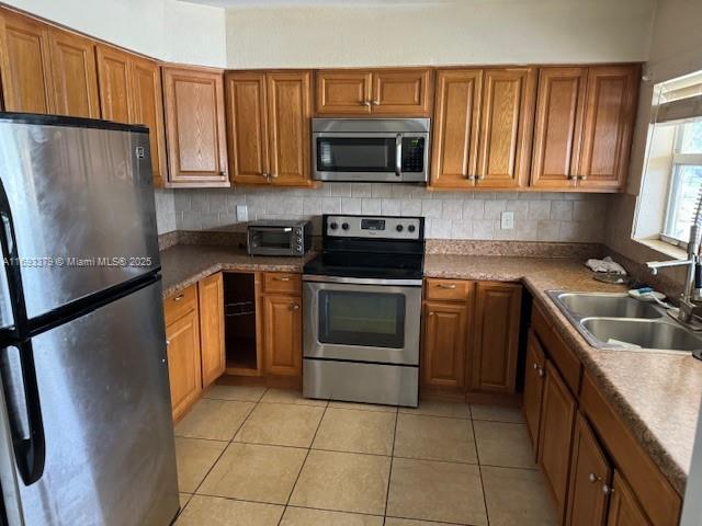 kitchen with appliances with stainless steel finishes, a sink, brown cabinetry, decorative backsplash, and light tile patterned floors