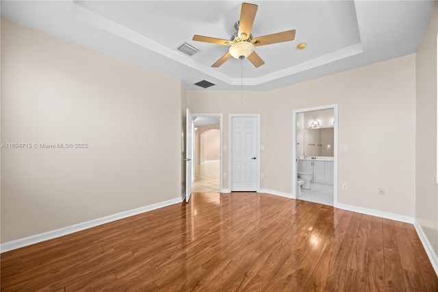 unfurnished bedroom with visible vents, baseboards, wood finished floors, ensuite bath, and a tray ceiling
