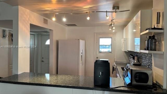 kitchen with backsplash, rail lighting, white refrigerator with ice dispenser, dark stone countertops, and white cabinets