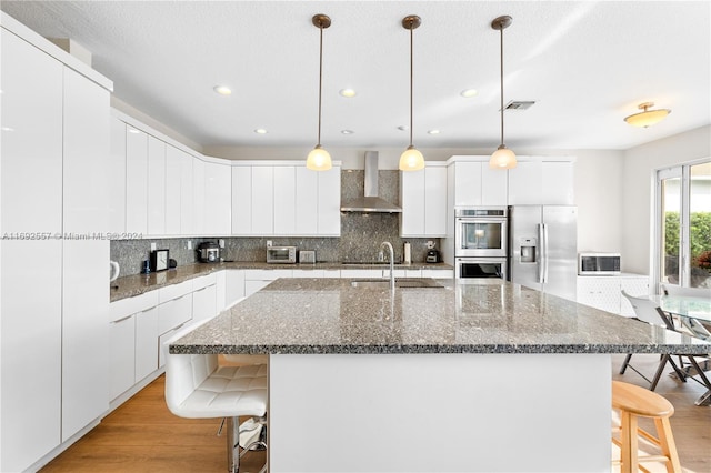 kitchen featuring appliances with stainless steel finishes, light wood-type flooring, wall chimney exhaust hood, white cabinetry, and an island with sink