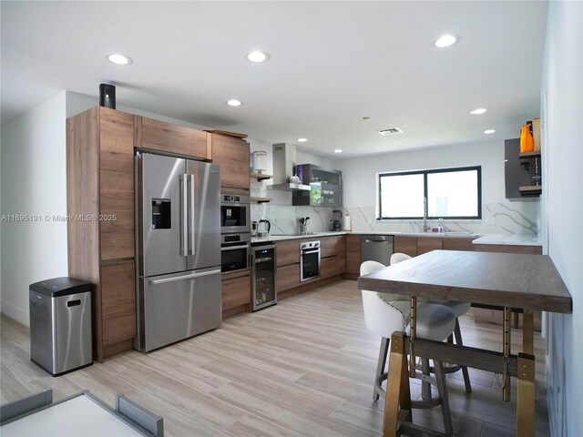 kitchen featuring wall chimney range hood, wine cooler, decorative backsplash, light wood-type flooring, and stainless steel appliances