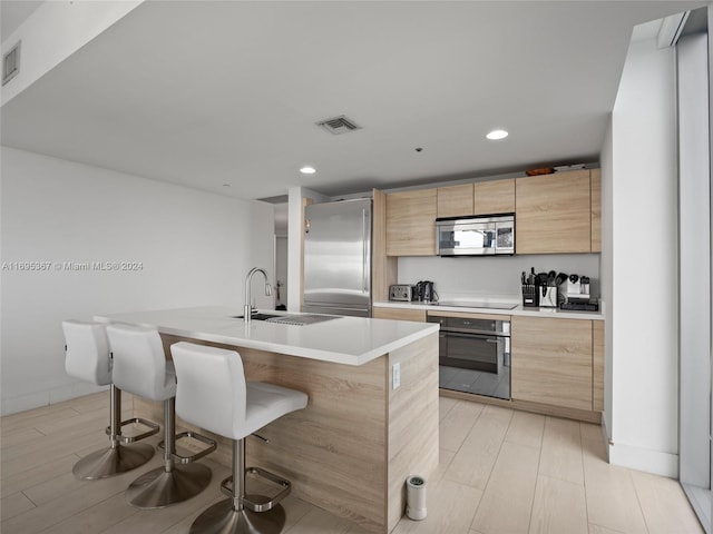 kitchen with sink, light brown cabinets, stainless steel appliances, a kitchen breakfast bar, and an island with sink