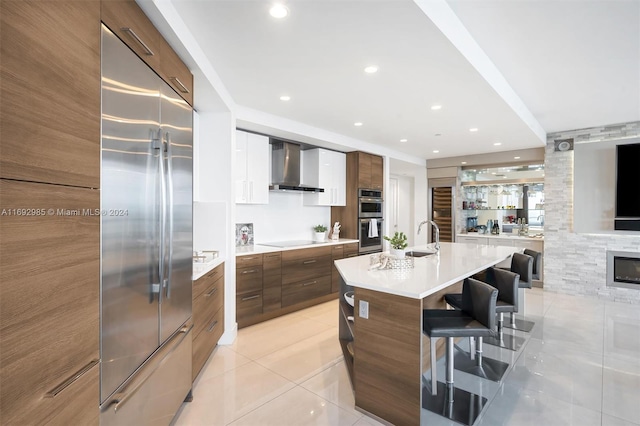 kitchen with a large fireplace, wall chimney exhaust hood, a kitchen bar, a center island with sink, and appliances with stainless steel finishes