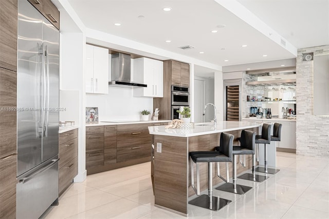 kitchen with wall chimney exhaust hood, a breakfast bar, stainless steel appliances, a center island with sink, and white cabinetry