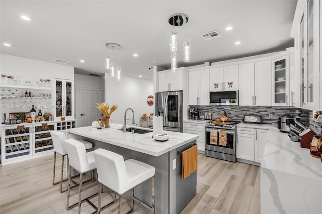 kitchen with white cabinetry, light stone counters, an island with sink, decorative light fixtures, and appliances with stainless steel finishes