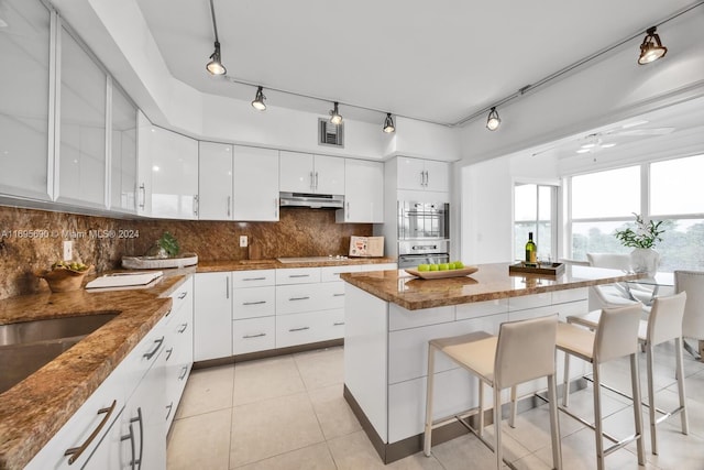 kitchen with decorative backsplash, stainless steel double oven, stone countertops, white cabinetry, and a breakfast bar area