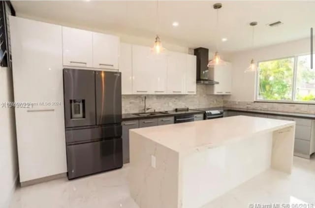 kitchen with wall chimney range hood, stainless steel fridge with ice dispenser, decorative light fixtures, a kitchen island, and white cabinetry