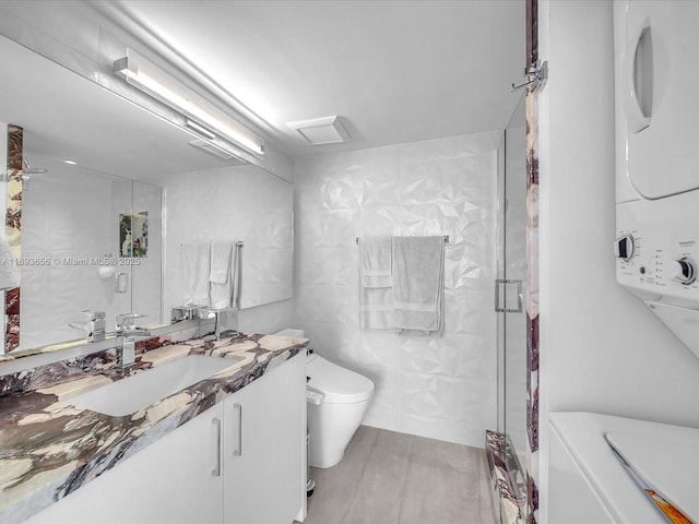 bathroom with toilet, stacked washer / dryer, tile walls, an enclosed shower, and vanity