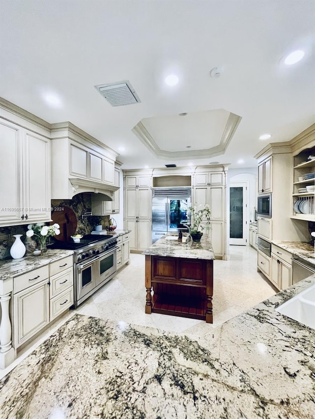 kitchen featuring a raised ceiling, crown molding, built in appliances, light stone countertops, and tasteful backsplash