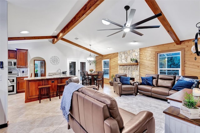 living room featuring ceiling fan with notable chandelier, wooden walls, light tile patterned floors, and vaulted ceiling with beams