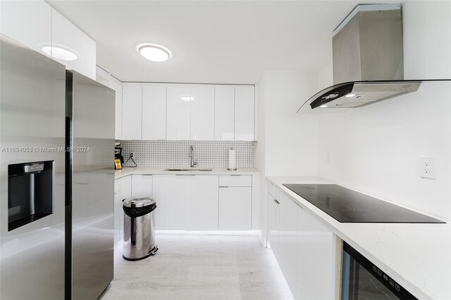 kitchen featuring wall chimney range hood, sink, black electric cooktop, light hardwood / wood-style floors, and white cabinetry