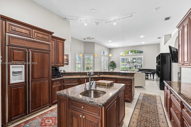 kitchen featuring paneled fridge, sink, decorative light fixtures, a center island with sink, and dark stone countertops