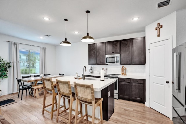 kitchen featuring an island with sink, pendant lighting, dark brown cabinets, appliances with stainless steel finishes, and light wood-type flooring