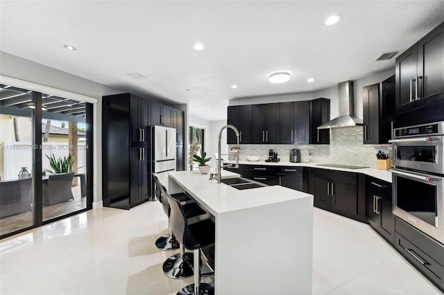 kitchen featuring sink, wall chimney exhaust hood, double oven, an island with sink, and black electric cooktop