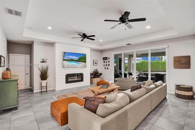 living room featuring ceiling fan, a large fireplace, and a tray ceiling