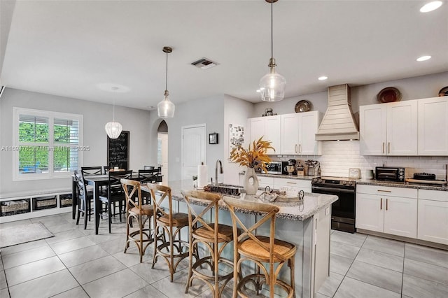 kitchen featuring premium range hood, an island with sink, pendant lighting, electric range oven, and a kitchen bar