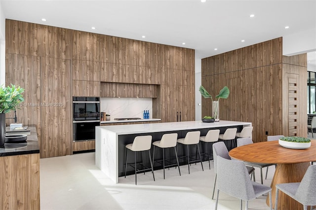 kitchen with black double oven, wooden walls, gas stovetop, a large island, and a kitchen bar