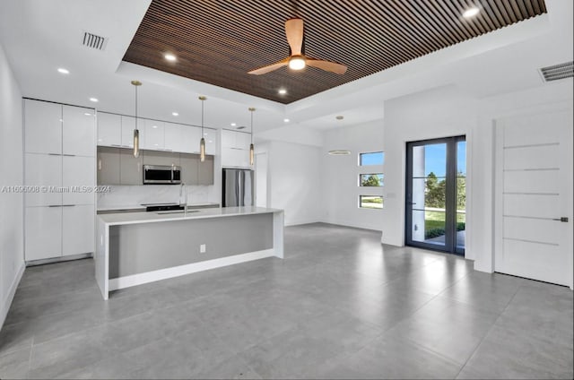 kitchen with stainless steel appliances, a raised ceiling, a kitchen island with sink, pendant lighting, and white cabinetry