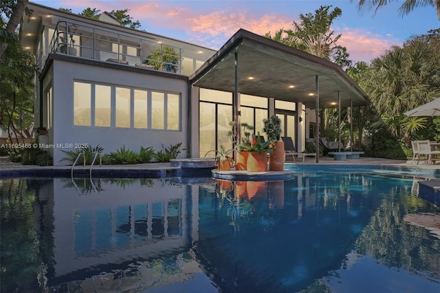 back of house at dusk with a balcony, an outdoor pool, a patio, and stucco siding