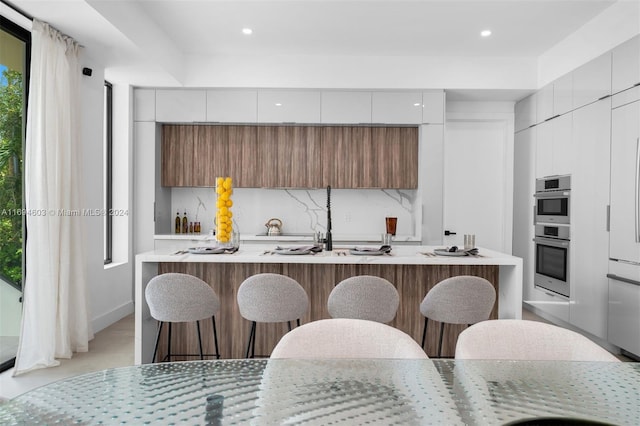 kitchen with a breakfast bar, white cabinetry, an island with sink, and double oven