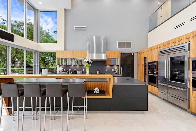 kitchen with a breakfast bar, exhaust hood, decorative backsplash, a towering ceiling, and appliances with stainless steel finishes