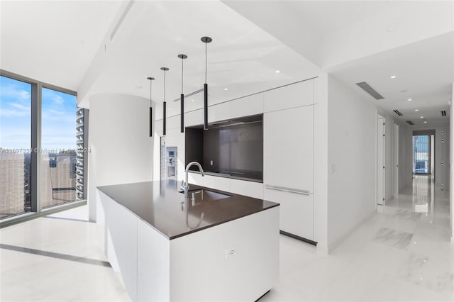 kitchen featuring floor to ceiling windows, a center island with sink, sink, hanging light fixtures, and white cabinetry