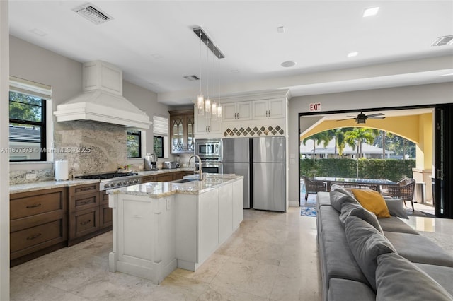 kitchen with premium range hood, appliances with stainless steel finishes, sink, hanging light fixtures, and a kitchen island with sink