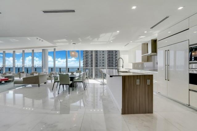 kitchen featuring wall chimney exhaust hood, a kitchen island with sink, pendant lighting, a wall of windows, and white cabinetry