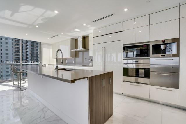 kitchen with sink, paneled fridge, wall chimney range hood, an island with sink, and white cabinets