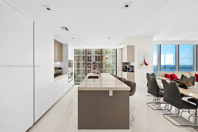 kitchen featuring sink, a wall of windows, an island with sink, white cabinetry, and light tile patterned flooring