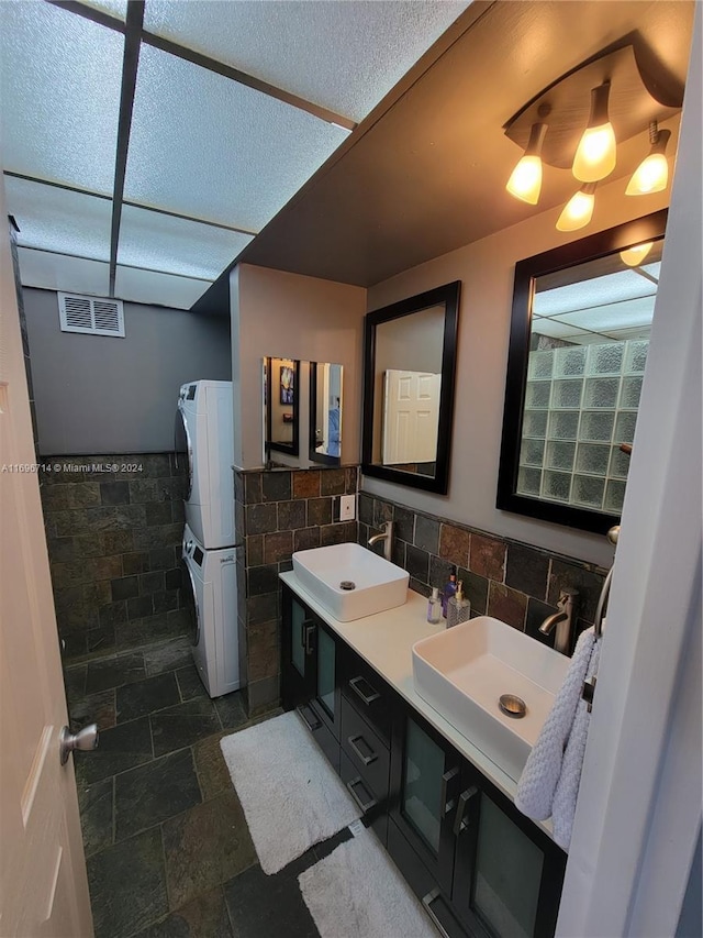 bathroom featuring tile walls, stacked washer and dryer, stone tile flooring, and a sink